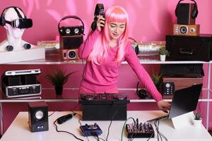 Dj with pink hair playing eletronic song at professional mixer console listening music using headset, enjoying nightlife. Asian perfomer having fun in club, dacing with fans during performance photo