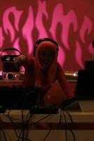 Artist with pink hair wearing headset while playing techno song at professional mixer console, enjoying night life in studio with pink background. Asian dj performing electronic music in club photo
