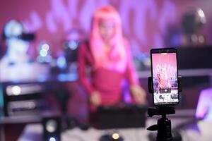 Artist with pink hair mixing song at professional turntables having fun while playing techno song in club at nightime. Dj woman recording music session with smartphone camera, enjoying nightlife photo