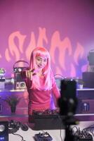 Woman with pink hair playing techno song at professional turntables while filimg process with mobile phone camera. Artist doing performance at nightclub with professional audio equipment photo