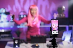 Woman with pink hair standing at dj table mixing song with turntables in front of camera while recording video with phone. Smiling artist doing performance at nightclub with audio equipment photo