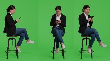 Modern female model browsing internet on smartphone, acting relaxed on chair in studio. Company employee in suit using mobile phone app to text messages, full body greenscreen backdrop. photo