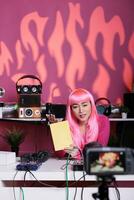 Influencer with pink hair presenting notebook in front of camera recording school supplies review using professional vlogging equipment. Asian vlogger filming podcast, live streaming photo