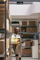 African american employee holding clipboard and checking inventory report in storage building. Storehouse manager making overview of factory products and looking at camera photo