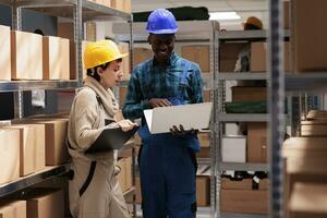 Man and woman warehouse coworkers supervising inventory together and reading report on laptop. Diverse employees working in stockroom and recording storehouse activity on computer photo