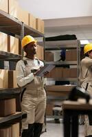 African american warehouse manager making freight overview in stockroom and checking inventory report on digital tablet. Storehouse operator managing cargo receiving and distribution photo