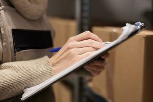 Postal office storage worker holding clipboard in warehouse, marking inventory checklist. Warehouse employee hand writing with pen close up, checking cardboard boxes in storehouse photo