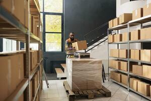 Employee wearing protective overall working in warehouse, carrying cardboard box and suitcase during small business inventory. African american supervisor preparing clients merchandise order photo