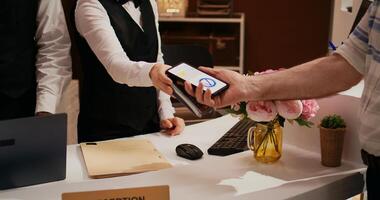 Elderly man makes nfc transaction with mobile phone at pos terminal, paying for accommodation at five star resort. Tourist arriving at front desk doing check in for holiday. Close up. Handheld shot. photo