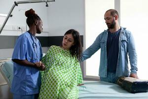 African american assistant helping pregnant woman to get out of bed in hospital ward, preparing patient for caesarean surgery. Future parents expecting child in maternity clinic photo