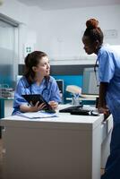 Nurse colleagues in happy relaxed hospital workspace working on tablet in modern professional medical office. Diverse healthcare specialists coworkers in modern clinic using technology photo