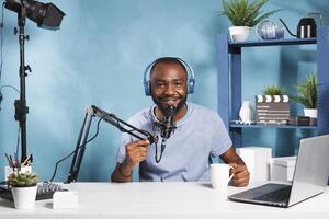 Smiling vlogger in headphones speaking into microphone while streaming on internet channel. Blogger wearing headset talking in mic, creating web content and recording podcast portrait photo