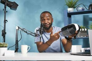 Blogger advertising virtual reality glasses for online blog and speaking in microphone. Web content creator showing virtual reality goggles and talking about metaverse on technology vlog photo