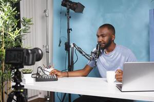 Vlogger using sound controller equipment and drinking coffee while recording video for internet channel. African american man creating digital content for blog with volume mixer photo