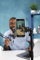 sonriente blogger en teléfono inteligente pantalla En Vivo transmisión en social medios de comunicación canal. joven alegre africano americano hombre creando digital contenido mientras grabación vídeo en móvil teléfono foto