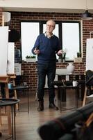 Senior man wearing glasses teaching art to adults in group class, standing in classroom introducing to drawing during creative workshop. Learning to draw from proffesional instructor photo