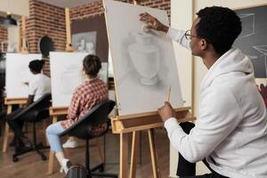 African American guy sitting at easel learning drawing techniques, sketching vase on canvas while attending creative group art class with friends. Creativity and leisure concept photo