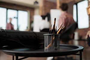 Sketching course. Close up of metal stand holder with graphite pencils standing on table against group of people students sitting at easels on blurred background, studying drawing in art studio photo