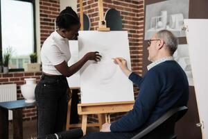 Diverse different aged art school students communicating during drawing lesson, looking at artwork on canvas and discussing sketching techniques. African American girl helping senior man at art class photo