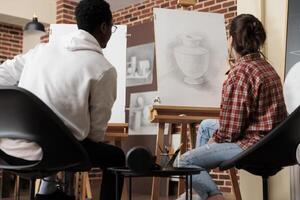 joven diverso personas estudiantes mirando a lona con florero bosquejo, desarrollar habilidades en línea y forma mientras estudiando en Arte academia. multirracial Pareja sentado a caballetes aprendizaje a dibujar juntos foto