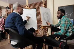 Two diverse men art school students talking during lesson, sitting at easels discussing artwork during creative group class, friends learning basics of drawing and sketching together photo