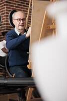 Concentrated focused senior man sitting at easel drawing object with graphite pencil. Mature student looking at ceramic vase making sketch on canvas during group art class in studio photo