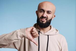 Arab man using thumb down gesture to show disapprovement while looking at camera with negative emotion grimace. Young adult person expressing disagreement studio portrait photo