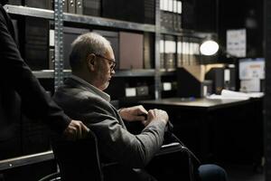 Paralyzed private detective wheelchair user and assisting coworker in criminal cases archive room. Senior investigator with disability surrounded by criminology folders on cabinet shelves photo