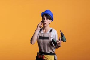 Surprised amazed builder talking on phone call and holding drilling gun, using smartphone in studio. Female contractor answering mobile phone for remote chat before renovating project. photo