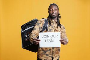 Takeaway employee holding job offer sign, recruiting people for food delivery in studio over yellow background. African american man hiring candidate for courier position. Advertising takeout career photo
