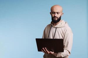 Focused young arab man holding laptop, analyzing code while developing software app. Confused person with puzzled expression thinking while using application on portable computer photo