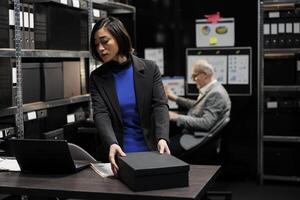 Professional asian accountant checking bureaucratic analytical data paperwork in file cabinet room office surrounded by accountancy statistic reports. Businesswoman in storage repository photo