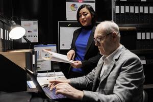 Professional business workers checking administrative analytical data in dark file room surrounded by accountancy statistic chart graphics. Bureaucratic office filled with invoice folders photo