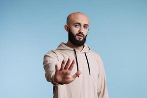 Confident arab man making stop gesture with hand, expressing rejection portrait. Young person in casual hoodie showing no signal with palm and looking at camera with serious emotions photo