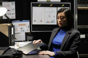 Asian businesswoman shutting down laptop at work preparing to go home. Bookkeeping consultant executive surrounded by accountancy file cabinet office statistics data flowcharts photo