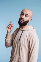 Inspired arab man having idea and raising index finger up while looking at camera. Young bald bearded person with excited facial expression pointing upwards studio portrait photo