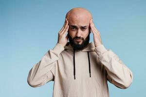 Exhausted arab man feeling tension in temples while suffering from headache studio portrait. Young bald bearded person having headache and looking at camera with pain grimace photo