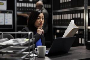 Bureaucratic administration employee checking accountancy budget plan data paperwork on laptop. Asian businesswoman executive in repository filled with document folders and graphical chart reports photo