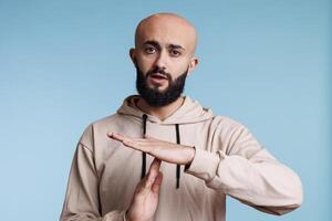 Arab man making time out gesture with hands while looking at camera. Young bald bearded person pausing conversation, showing interruption signal with arms studio portrait photo