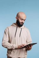 Young arab man using application on digital tablet, typing message on touchscreen. Focused person holding portable gadget, browsing internet pages and scrolling social media photo
