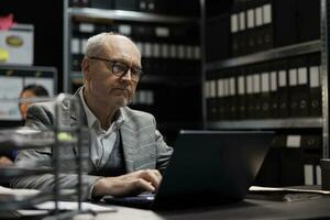 Senior private detective checking confidential criminology evidence files on laptop. Investigator in agency file storage office surrounded by folders on cabinet shelves photo