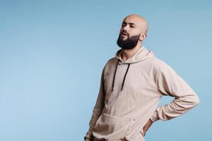 Young arab man suffering from chronic lower back pain while rubbing sore muscles. Arabian person wearing casual hoodie clothes grimacing while having backache and spine problems photo