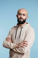 Confident arab man standing with crossed arms while looking at camera. Young serious arabian person wearing casual hoodie clothes posing with folded hands studio portrait photo