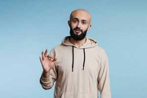 Relaxed arab man showing approval with ok symbol while looking at camera. Calm young bearded person making okay acceptance gesture with fingers while posing for studio portrait photo