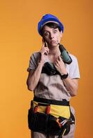 Bored construction worker answering smartphone call, posing over yellow background. Woman working as professional builder with power drilling gun, talking on mobile phone line. photo