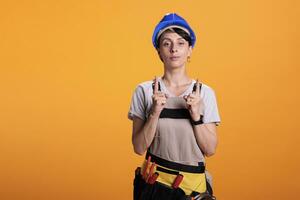 Portrait of woman builder pointing upwards with index fingers, showing above and indicating direction up. Posing in studio over yellow background and wearing hardhat with overalls. photo