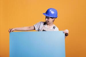 Construction worker holding empty cardboard sign for advertisement in studio. Optimistic woman renovator in uniform showing blank carton banner with isolated copyspace, mockup. photo