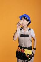 Construction worker serving coffee cup after renovation, using building tools to restore and decorate. Professional contractor wearing protective helmet and overalls in studio against yellow photo