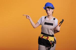 mujer constructor indicando dirección en izquierda lado de estudio, posando con par de alicates y señalando oblicuo. hembra construcción trabajador mirando a cámara y demostración lados, amarillo antecedentes. foto