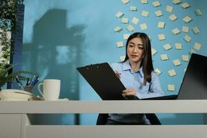 Corporate asian employee using laptop to do an online research and taking notes on clipboard. Businesswoman sitting at office desk concentrating on taking notes filling out form. photo